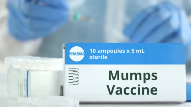 Ampuller med påssjuka vaccin på bordet nära fungerande laboratorieassistent, fiktiv logotyp på lådan — Stockvideo