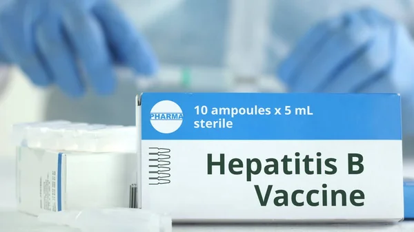 Schachtel mit Hepatitis-B-Impfstoff auf dem Tisch gegen verschwommene Laborassistentin. Fiktives pharmazeutisches Logo — Stockfoto