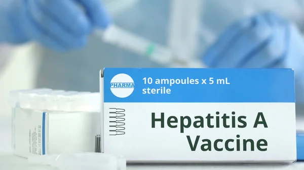 Kartong med hepatit A vaccin på bordet mot suddig labbassistent eller läkare. Fiktiva läkemedlets logotyp — Stockfoto