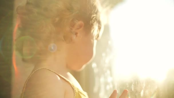 Imádnivaló göndör szőke kislány néz ki az ablakon otthon és mosolyog a kamerába — Stock videók