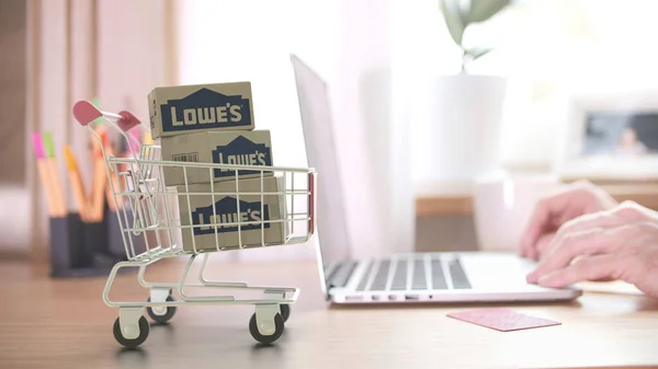 Dobozok LOWES logóval a bevásárlókocsiban a vásárló közelében laptoppal. Szerkesztői online vásárlás otthonról 3D renderelés — Stock Fotó