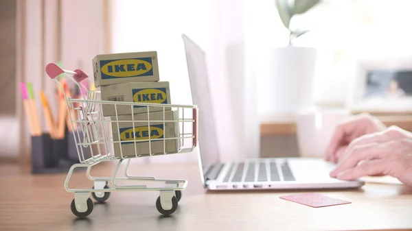 Három kartondoboz IKEA logóval a bevásárlókocsiban a vásárló közelében laptoppal. Szerkesztői online vásárlás otthonról 3D renderelés — Stock Fotó