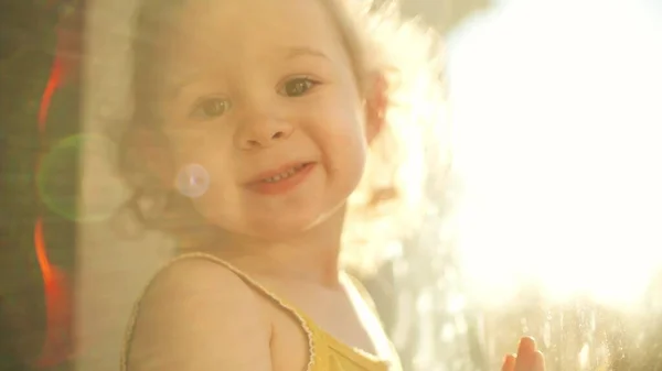 Rozkošné kudrnaté blondýny holčička se usmívá pro kameru v blízkosti okna — Stock fotografie