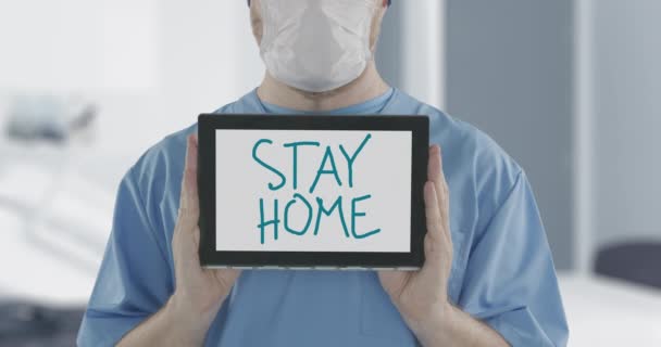 戴口罩的医生拿着平板电脑，上面有"待在家里"的手写文字。COVID-19疾病爆发相关夹期间的自我隔离 — 图库视频影像