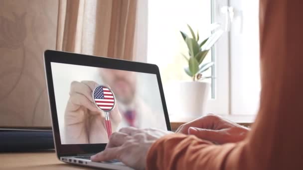 医者のビデオ通話は自宅のノートパソコンにアメリカ国旗のついた聴診器のベル。アメリカのテレメディシン — ストック動画