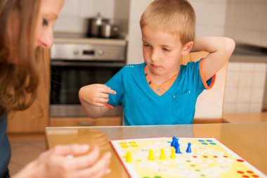 young boy plays ludo game with his mother on a table in livingroom clipart