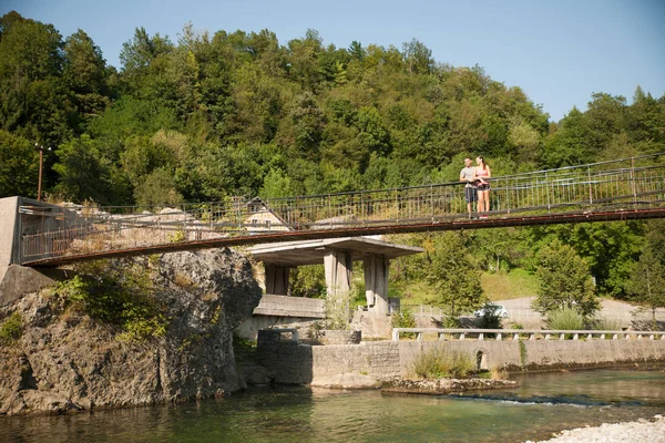 Giovane coppia attiva su un ponte sul fiume alpino in estate calda — Foto Stock