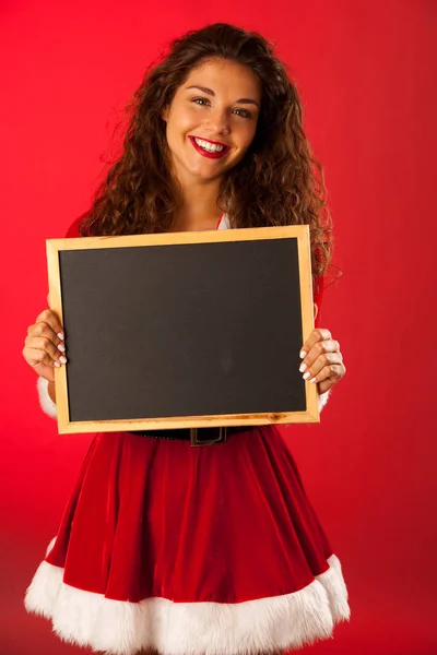 Bela jovem mulher em santa dres detém blackboard sobre vermelho ba — Fotografia de Stock