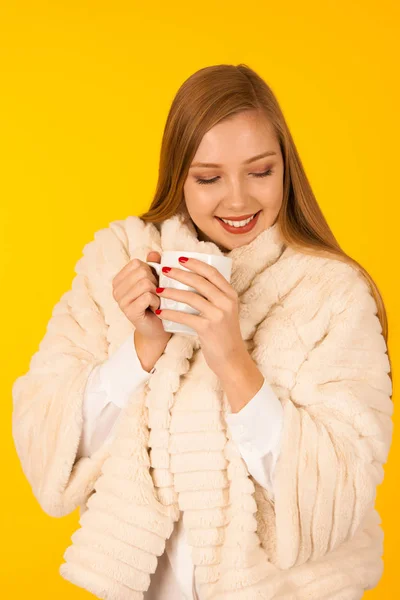Beautiful woman in a fur winter jacket drinks tea over yellow ba — Stock Photo, Image