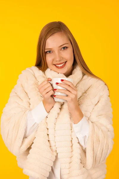 Beautiful woman in a fur winter jacket drinks tea over yellow ba — Stock Photo, Image