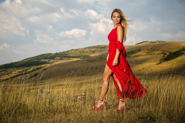 夏の赤いドレスの美しいブロンドが山のフィールドを歩いています — ストック写真
