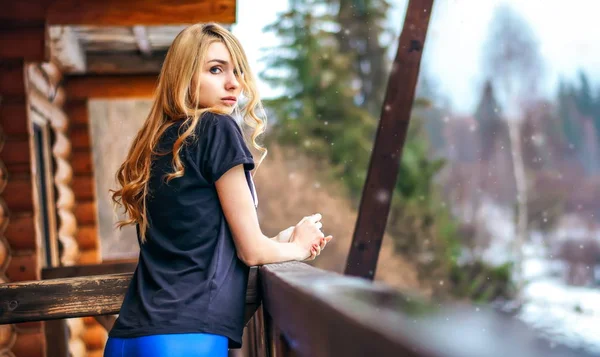 Mooi meisje permanent op de veranda van een houten huis in de winter bos en lome teder kijkt — Stockfoto
