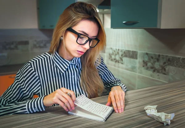 girl in glasses reads and looks at records in a paper notebook sitting at the table at home