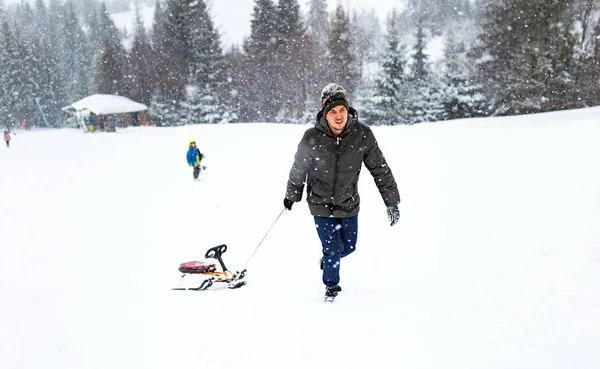 Homem puxa trenó na neve, floresta de inverno flocos de neve queda de neve, jogar atividade divertida — Fotografia de Stock