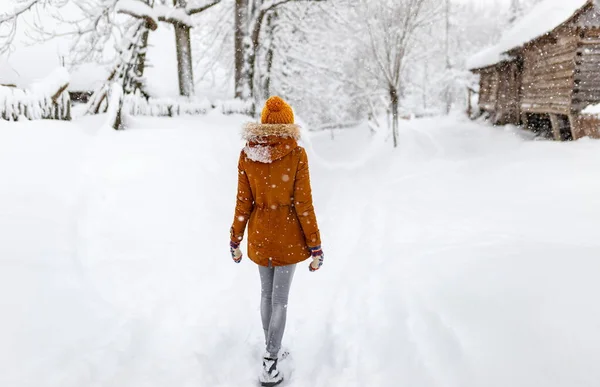 女の子は冬に戻り田舎の吹雪の雪の外の白い村の雪の寒さ、黄色の帽子の旅行を歩く ロイヤリティフリーのストック画像