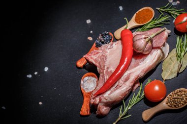 Kaşıklar, biberler, sarımsak, defne yaprağı, acı biber domatesleri ve siyah bir yüzey üzerinde büyük ve taze bir parça et.