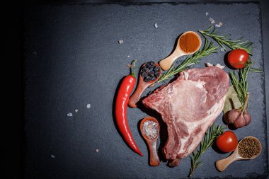 Kaşıklar, biberler, sarımsak, defne yaprağı, acı biber domatesleri ve siyah bir yüzey üzerinde büyük ve taze bir parça et.