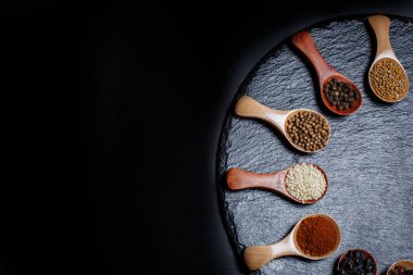 Farklı baharatlar, biberler, kişniş, susam tohumları ve yemek pişirmek için diğerleri siyah arka planda tahta kaşıklarda.
