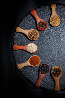 Farklı baharatlar, biberler, kişniş, susam tohumları ve yemek pişirmek için diğerleri siyah arka planda tahta kaşıklarda.