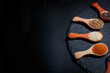 Farklı baharatlar, biberler, kişniş, susam tohumları ve yemek pişirmek için diğerleri siyah arka planda tahta kaşıklarda.