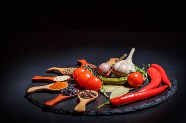 Ahşap kaşıklarda baharatlar ve kiraz domatesleri, kırmızı biber, sarımsak ve siyah bir zeminde pişirmek için şifalı otlar.