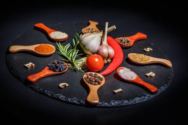 Ahşap kaşıklarda baharatlar ve kiraz domatesleri, kırmızı biber, sarımsak ve siyah bir zeminde pişirmek için şifalı otlar.