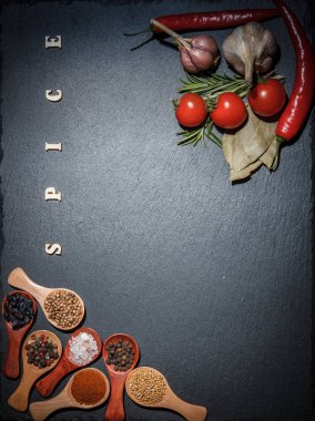 Ahşap kaşıklarda baharatlar ve kiraz domatesleri, kırmızı biber, sarımsak ve siyah bir zeminde pişirmek için şifalı otlar.