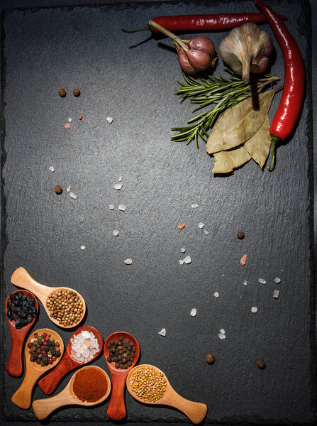 Spices in wooden spoons and cherry tomatoes, chili peppers, garlic and herbs for cooking on a black surface and black background