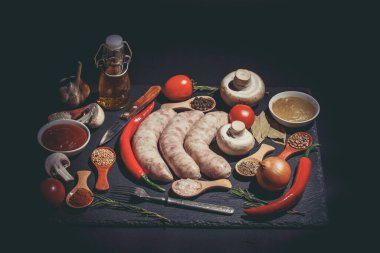 Çiğ sosis, farklı sebzeler, baharatlar ve siyah arka plandaki malzemeler.