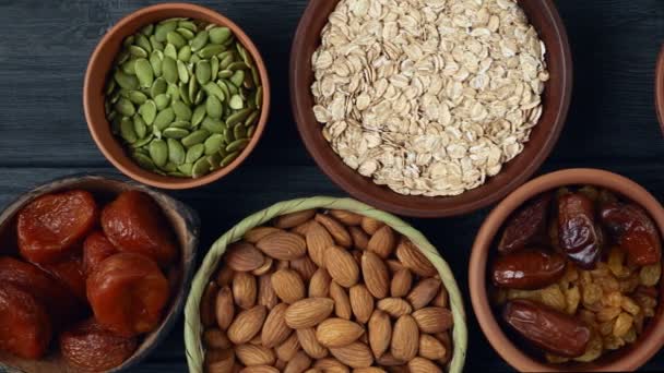 Desayuno saludable. Granola. Harina de avena, copos de cebada. Nueces: avellanas, almendras, anacardos. Frutas secas: pasas, dátiles, albaricoques secos. En tazones de barro sobre un fondo negro de madera. Vista de cámara lenta.Vista superior . — Vídeos de Stock