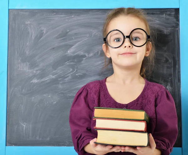 Gadis kecil lucu berdiri di dekat papan tulis dengan kacamata besar yang lucu. — Stok Foto