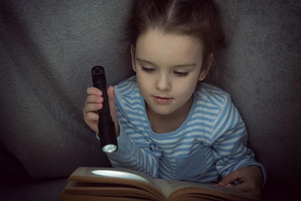 Niña leyendo cuentos de hadas libro bajo las cubiertas en la víspera — Foto de Stock