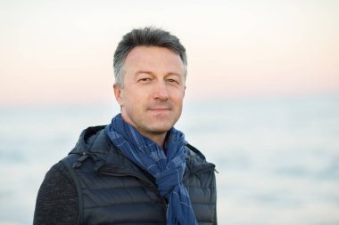 Kumsaldaki yakışıklı, orta yaşlı adam. Çekici, mutlu, orta yaşlı bir erkek model. Gün batımında deniz kenarında poz veriyor. Gün doğumunda. Güzel bir adamın açık hava portresi.. 