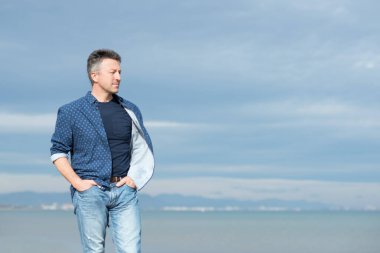 Kumsalda yürüyen orta yaşlı yakışıklı bir adam. Sahilde kot pantolon ve tişörtle poz veren çekici orta yaşlı erkek manken. Güzel maço bir adamın açık hava portresi.. 