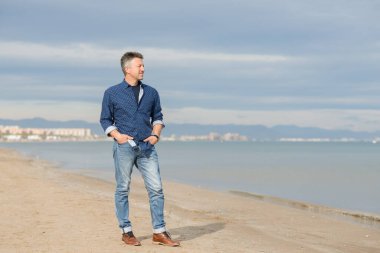 Kumsalda yürüyen orta yaşlı yakışıklı bir adam. Çekici, mutlu, orta yaşlı erkek manken deniz kenarında kot pantolon ve tişörtle poz veriyor. Güzel, maço bir adamın açık hava portresi.. 
