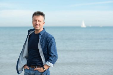 Kumsalda yürüyen orta yaşlı yakışıklı bir adam. Sahilde kot pantolon ve tişörtle poz veren çekici orta yaşlı erkek manken. Güzel maço bir adamın açık hava portresi.. 