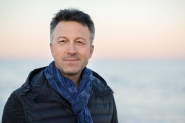 Kumsaldaki yakışıklı, orta yaşlı adam. Çekici, mutlu, orta yaşlı bir erkek model. Gün batımında deniz kenarında poz veriyor. Gün doğumunda. Güzel bir adamın açık hava portresi.. 