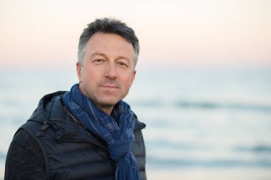 Kumsaldaki yakışıklı, orta yaşlı adam. Çekici, mutlu, orta yaşlı bir erkek model. Gün batımında deniz kenarında poz veriyor. Gün doğumunda. Güzel bir adamın açık hava portresi.. 