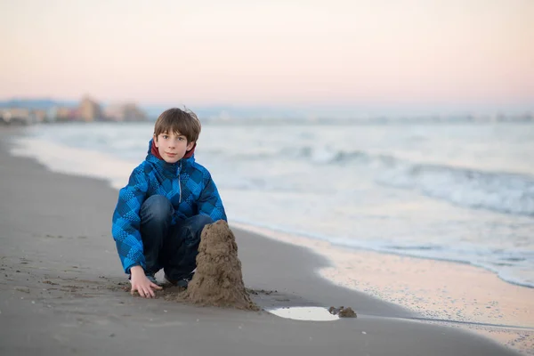 若い男の子は冬のビーチで砂の城を構築します かわいい11歳の少年海辺で 夜の時間 子供の屋外肖像画 — ストック写真