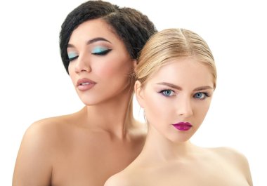 Makyaj. Çifte kadın portresi. Kafkasyalı sarışın kız ve stüdyoda beyaz arka planda poz veren güzel melez genç kadın. Moda, güzellik, neşe, gençlik.