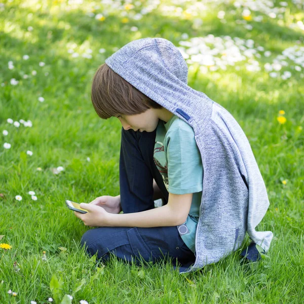 春のグレードでスマートフォンを使っている少年 春の公園で緑の芝生に座ってかわいい11歳の少年 子供の屋外肖像画 — ストック写真