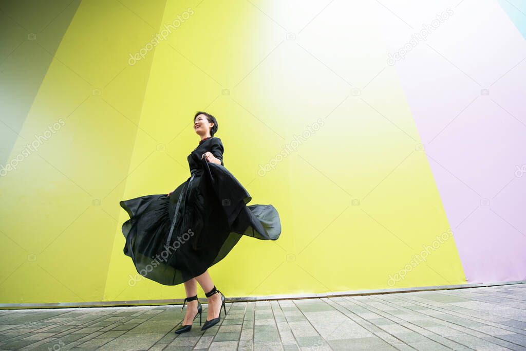 Fashion woman. Young beautiful chinese girl dancing outdoor wearing long black dress with high heels over colorful wall background. Stylish trendy lady. 