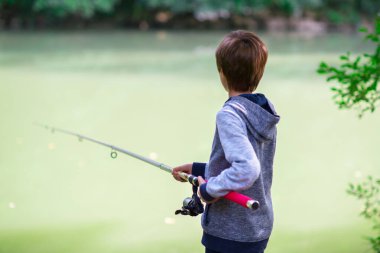 Genç balıkçı. Nehir kıyısında balık tutan bir çocuk, yazın açık havada. Genç çocuk elinde olta ve balıkla nehir kıyısında oturuyor. Yaz tatili aktivitesi. Balık tutmaya. Açı.