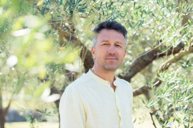 Zeytin ağaçları. Zeytin bahçesinde poz veren yakışıklı bir adam. Akdeniz zeytin tarlasında hasat için hazır erkek portreleri. İtalyan zeytinliğinde kendine güvenen olgun bir adam taze zeytinlerle dolu. Taze zeytin. Zeytin çiftliği