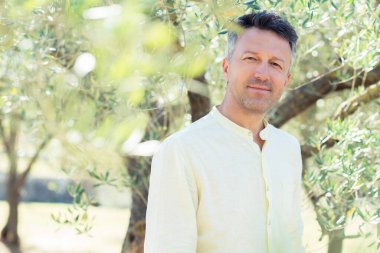 Zeytin ağaçları. Zeytin bahçesinde poz veren yakışıklı bir adam. Akdeniz zeytin tarlasında hasat için hazır erkek portreleri. İtalyan zeytinliğinde kendine güvenen olgun bir adam taze zeytinlerle dolu. Taze zeytin. Zeytin çiftliği