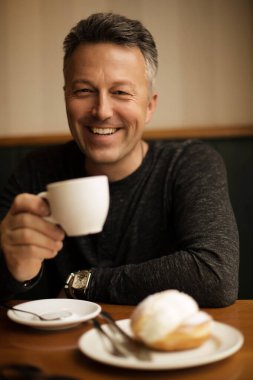 Çekici gülümseyen, orta yaşlı bir adam kafede sabah kahvesini içiyor. Görüntü tonlu. Kafedeki yakışıklı bekar adam.