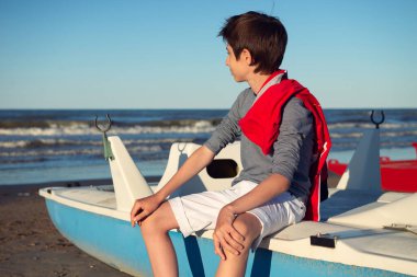 Yaz plajında katamaranın üzerinde oturan genç bir çocuk. Sevimli gözlüklü, gülümseyen mutlu 12 yaşında deniz kenarında denize bakan bir çocuk. Çocuğun deniz kenarındaki açık hava portresi..