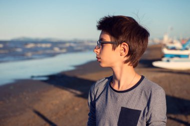 Yaz plajında poz veren genç bir çocuk. Sevimli sakin gözlüklü, deniz kenarında 12 yaşında bir çocuk, denizde lokma yiyor. Profilde çocuğun açık hava portresi.