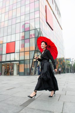 Pekin 'in merkezinde yürüyen moda kadını. Dışarıda poz veren genç, güzel Çinli bir kız uzun siyah bir elbise ve yüksek topuklu ayakkabıyla renkli duvar arkasında kırmızı bir şemsiye tutuyor. Modaya uygun bir bayan..