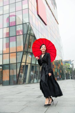 Pekin 'in merkezinde yürüyen moda kadını. Dışarıda poz veren genç, güzel Çinli bir kız uzun siyah bir elbise ve yüksek topuklu ayakkabıyla renkli duvar arkasında kırmızı bir şemsiye tutuyor. Modaya uygun bir bayan..